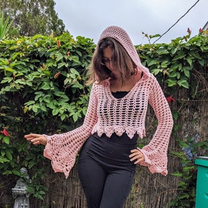 May include: A light pink, open-weave crochet hooded top with bell sleeves. The top has a scalloped hem and is worn over a black top. The model is wearing black pants. The background includes green foliage and a cloudy sky.