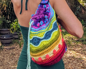 May include: A woman wearing a green dress with a crocheted rainbow drawstring bag. The bag has a variety of colours, including purple, blue, green, yellow, orange, and red.