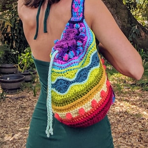 May include: A woman wearing a green dress with a crocheted rainbow drawstring bag. The bag has a variety of colours, including purple, blue, green, yellow, orange, and red.