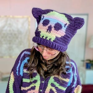 May include: A dark purple crochet cardigan featuring a multi-coloured skeleton rib cage design in pastel hues of pink, blue, and green. The matching dark purple crochet cat-ear beanie displays a pastel skull graphic on the front.
