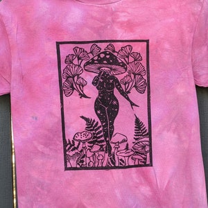 May include: A pink tie-dye t-shirt with a black and white print of a woman standing under a mushroom. The woman is surrounded by mushrooms and leaves. The print is in a rectangular frame.