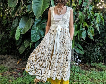 May include: A white crochet maxi dress with a tie waist and a scalloped hem. The dress is worn over a yellow skirt and the model is standing in a grassy area.