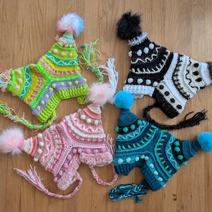 Puede incluir: Cuatro gorros de ganchillo hechos a mano con orejeras y pompones. Los gorros presentan intrincados patrones en varios colores, incluyendo verde, rosa, turquesa y blanco y negro. Cada gorro tiene un pompón esponjoso y borlas decorativas.