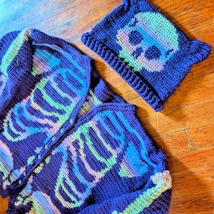 Op de afbeelding: Een donkerpaars gehaakt vest en een bijpassende muts met kattenoren. Beide items hebben een veelkleurig skelet-borstkas en schedelontwerp in tinten lichtgroen, blauw en roze. Het vest is voorzien van een knoopsluiting aan de voorzijde.