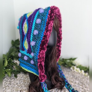 May include: A colorful crocheted hooded scarf with a purple, blue, and green geometric pattern. The scarf has a long, braided tassel and comes with written instructions and a video tutorial.
