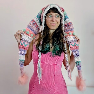 Puede incluir: Una capucha de ganchillo larga y multicolor con rayas horizontales en tonos rosa, morado, verde azulado y blanco. La capucha incluye detalles de puntos texturizados, lazos laterales trenzados y grandes pompones rosas esponjosos en los extremos.