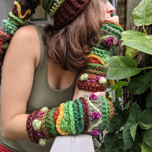 May include: A woman wearing a brown, green and yellow crocheted hat with a long, crocheted tail. She is also wearing matching crocheted fingerless gloves.