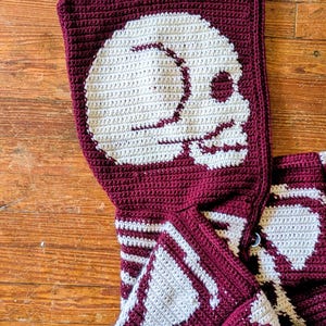 May include: A burgundy and white crochet hooded scarf featuring a large skull design on the hood. The skull is white, and the scarf has a button closure. The scarf is made of a soft, textured yarn.
