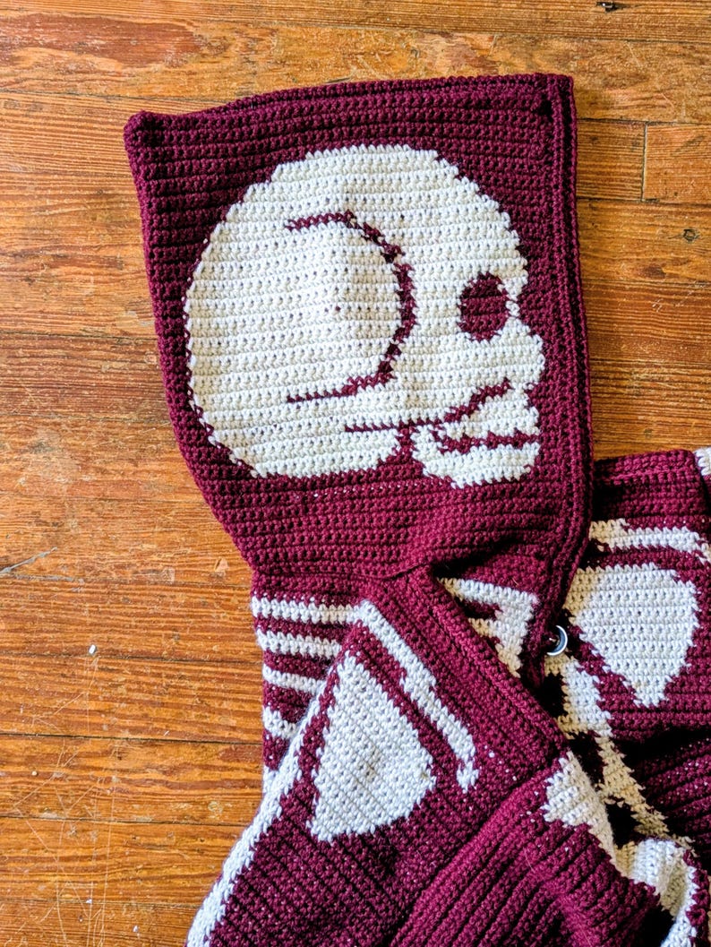 May include: A burgundy and white crochet hooded scarf featuring a large skull design on the hood. The skull is white, and the scarf has a button closure. The scarf is made of a soft, textured yarn.