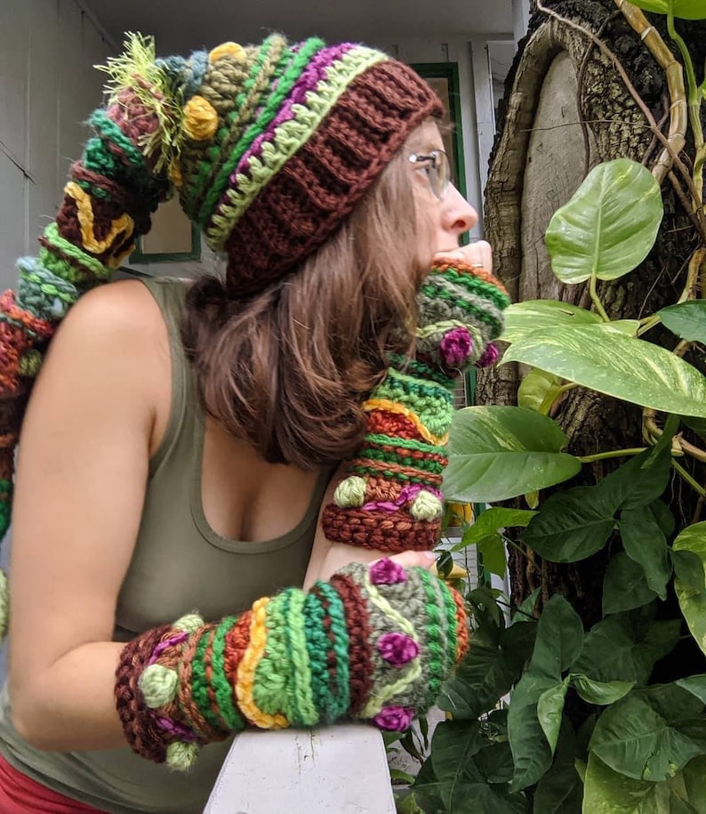 May include: A woman wearing a brown, green and yellow crocheted hat with a long, crocheted tail. She is also wearing matching crocheted fingerless gloves.