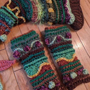 May include: A pair of crocheted fingerless gloves in a green, brown and purple colour scheme. The gloves feature a variety of textures and embellishments, including pom-poms and velvet ribbon.