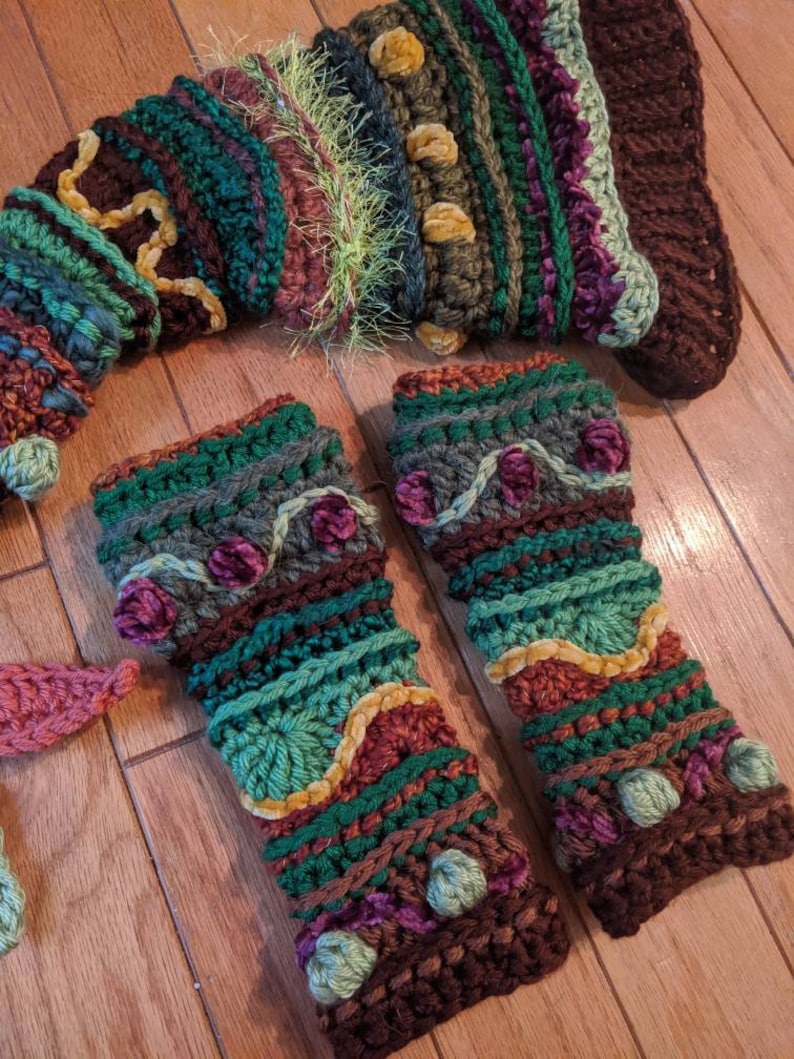 May include: A pair of crocheted fingerless gloves in a green, brown and purple colour scheme. The gloves feature a variety of textures and embellishments, including pom-poms and velvet ribbon.