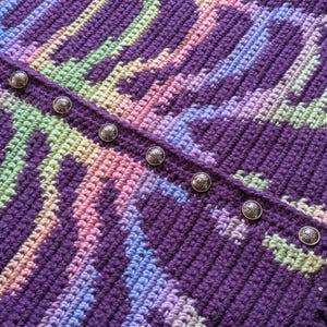 May include: Close-up of a handmade crochet garment in shades of purple, pink, blue, and green. The intricate pattern is accented by a row of decorative silver buttons. The textured fabric suggests a cozy, handcrafted item.