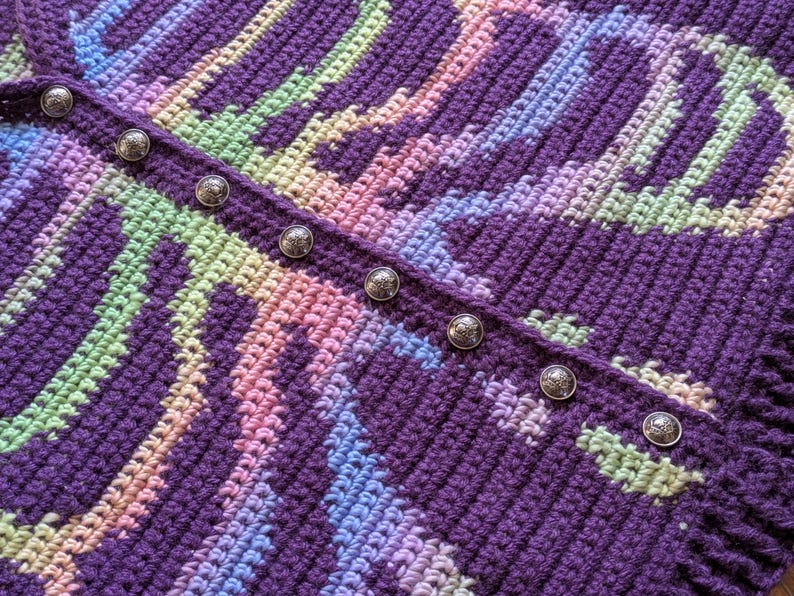 May include: Close-up of a handmade crochet garment in shades of purple, pink, blue, and green. The intricate pattern is accented by a row of decorative silver buttons. The textured fabric suggests a cozy, handcrafted item.