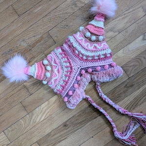 May include: A handmade, colorful crochet hat in shades of pink, white, and brown. The hat features a unique design with textured patterns, small pom-poms, and two large fluffy pink pom-poms. Braided ties with tassels complete the design.