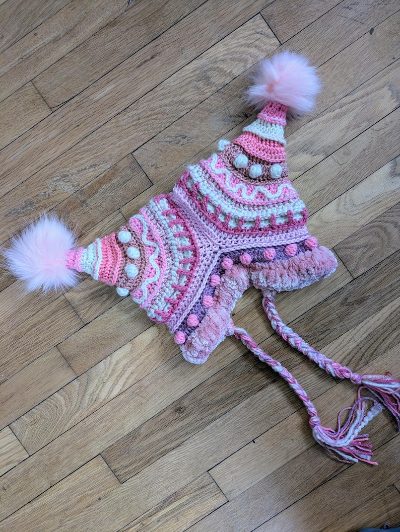 May include: A handmade, colorful crochet hat in shades of pink, white, and brown. The hat features a unique design with textured patterns, small pom-poms, and two large fluffy pink pom-poms. Braided ties with tassels complete the design.