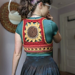 Puede incluir: Un chaleco de ganchillo con forma de girasol con un esquema de color rojo, naranja, amarillo y marr&oacute;n. El chaleco se lleva sobre una camisa turquesa.