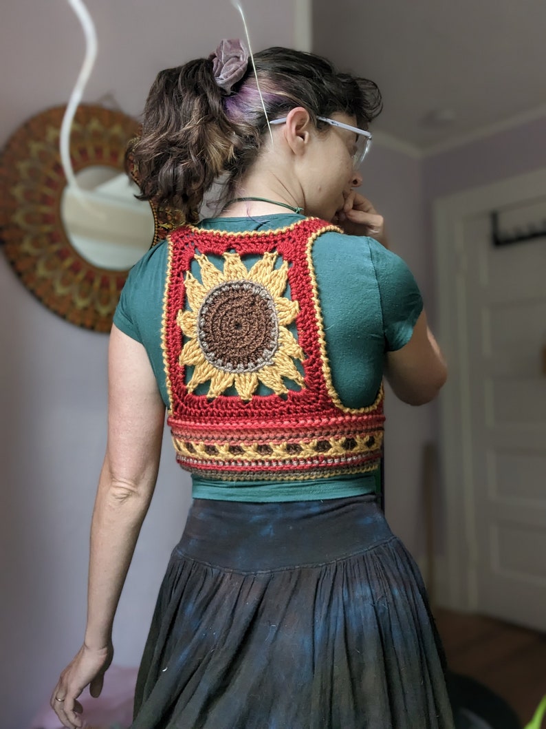Puede incluir: Un chaleco de ganchillo con forma de girasol con un esquema de color rojo, naranja, amarillo y marr&oacute;n. El chaleco se lleva sobre una camisa turquesa.