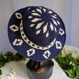 May include: A navy blue crochet sun hat with a wide brim. The hat features a cream-colored floral pattern around the crown and brim. A dark blue ribbon is sewn around the base of the crown. The hat is displayed on a black stand.