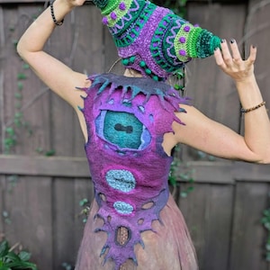 May include: A handmade, whimsical outfit featuring a cone-shaped hat and a vest. The hat is green and purple with intricate crochet details. The vest is purple with a large eye design and other circular accents. The outfit is likely for costume or fantasy wear.