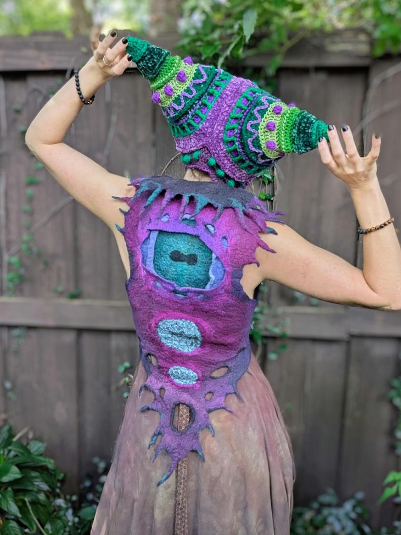 May include: A handmade, whimsical outfit featuring a cone-shaped hat and a vest. The hat is green and purple with intricate crochet details. The vest is purple with a large eye design and other circular accents. The outfit is likely for costume or fantasy wear.