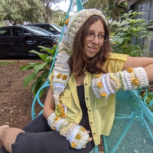 May include: A person wearing a crocheted hat and matching fingerless gloves with a floral design. The hat has a long, pointed hood and the gloves have yellow flowers. The person is sitting on a blue metal bench.