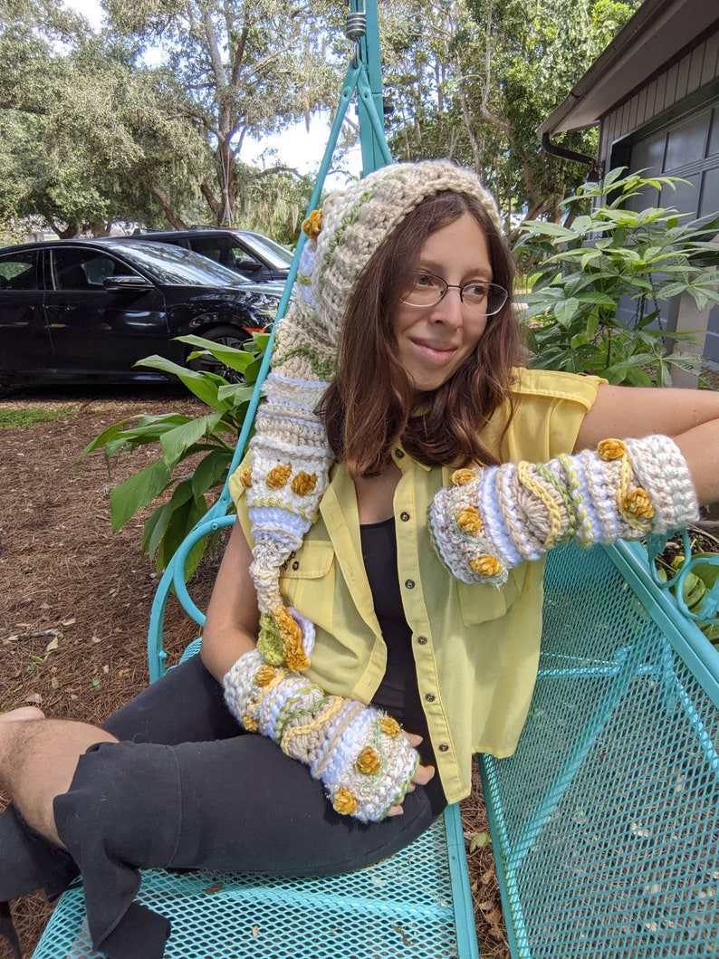 May include: A person wearing a crocheted hat and matching fingerless gloves with a floral design. The hat has a long, pointed hood and the gloves have yellow flowers. The person is sitting on a blue metal bench.