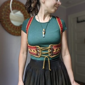Puede incluir: Una mujer con una camisa verde y un cors&eacute; de crochet rojo, marr&oacute;n y dorado. El cors&eacute; tiene cordones marrones y est&aacute; atado en la parte delantera. La mujer tambi&eacute;n lleva un collar con un colgante de cristal.