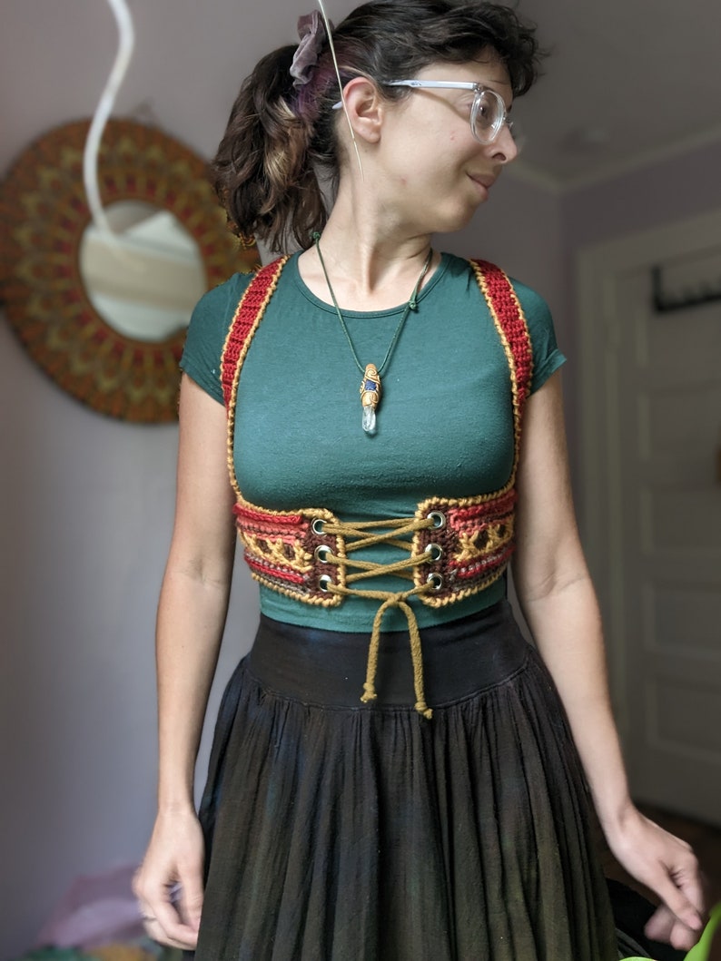 Puede incluir: Una mujer con una camisa verde y un cors&eacute; de crochet rojo, marr&oacute;n y dorado. El cors&eacute; tiene cordones marrones y est&aacute; atado en la parte delantera. La mujer tambi&eacute;n lleva un collar con un colgante de cristal.