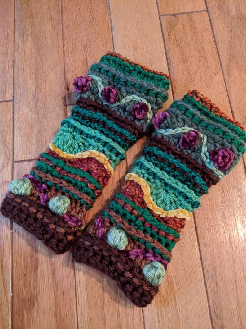 May include: A pair of brown, green and purple crocheted arm warmers with a floral design. The arm warmers have a textured, multi-coloured pattern with small pom-poms.