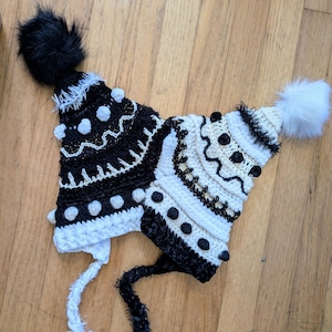 May include: Two handmade crochet hats. One is black and white with a black pom-pom and the other is white and black with a white pom-pom. Both hats have decorative bobbles and long ties.