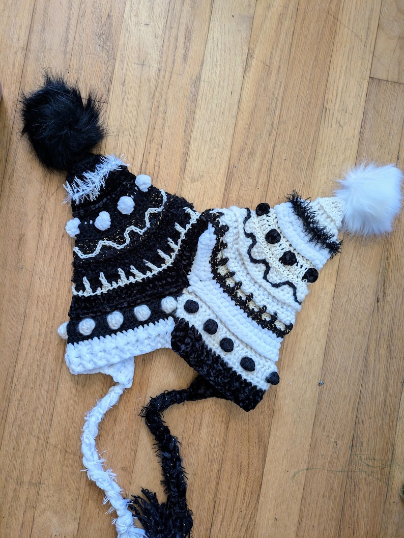 May include: Two handmade crochet hats. One is black and white with a black pom-pom and the other is white and black with a white pom-pom. Both hats have decorative bobbles and long ties.