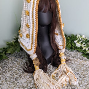 May include: White and yellow crocheted hooded scarf with tassels and gold accents. The scarf is worn on a mannequin with brown hair.