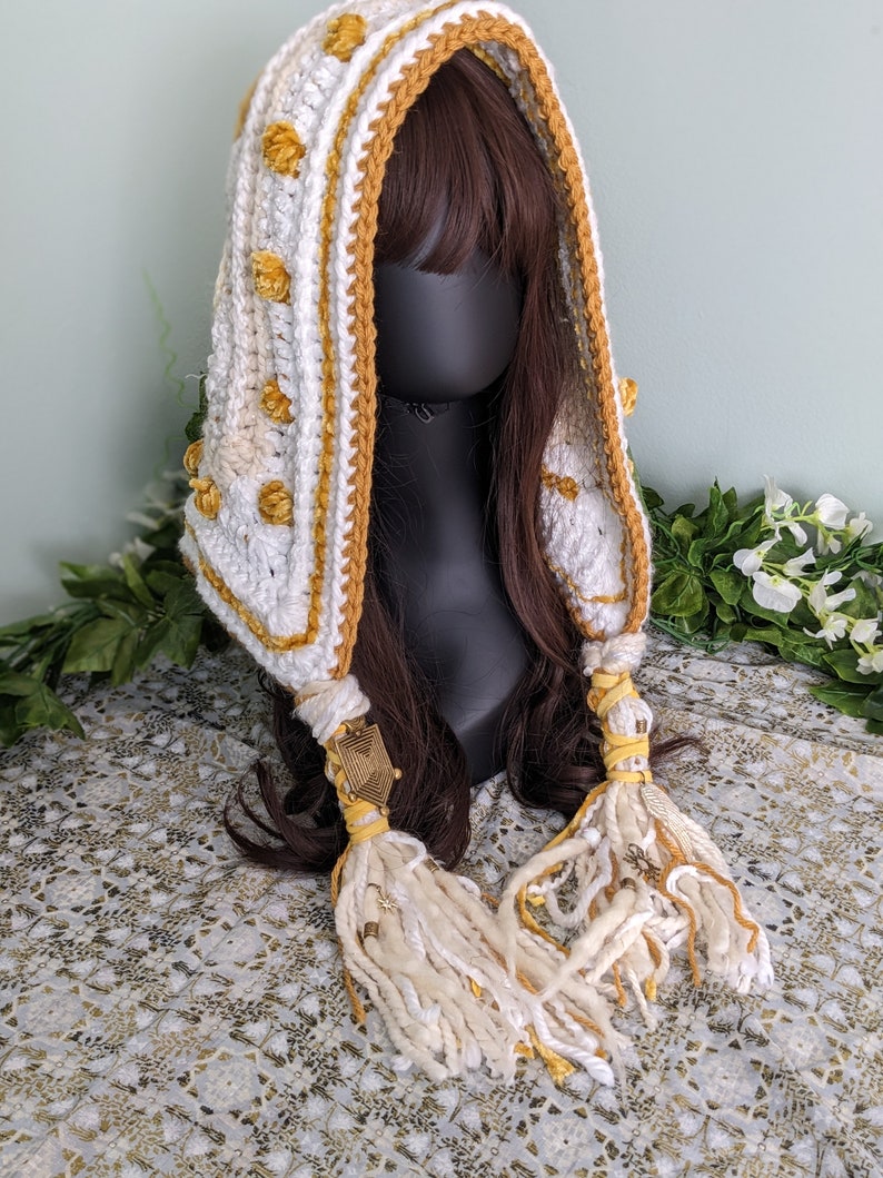 May include: White and yellow crocheted hooded scarf with tassels and gold accents. The scarf is worn on a mannequin with brown hair.