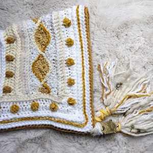 May include: White and gold crocheted hooded scarf with a geometric pattern and tassels. The scarf has a gold trim and a decorative metal charm.