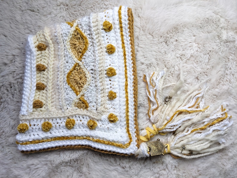 May include: White and gold crocheted hooded scarf with a geometric pattern and tassels. The scarf has a gold trim and a decorative metal charm.