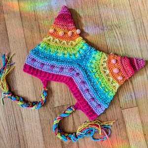 May include: A colorful, handmade crochet hat in a star shape. The hat features a rainbow of colors, including pink, orange, yellow, green, and blue. It has braided ties with colorful tassels. The hat is likely for a child or adult.