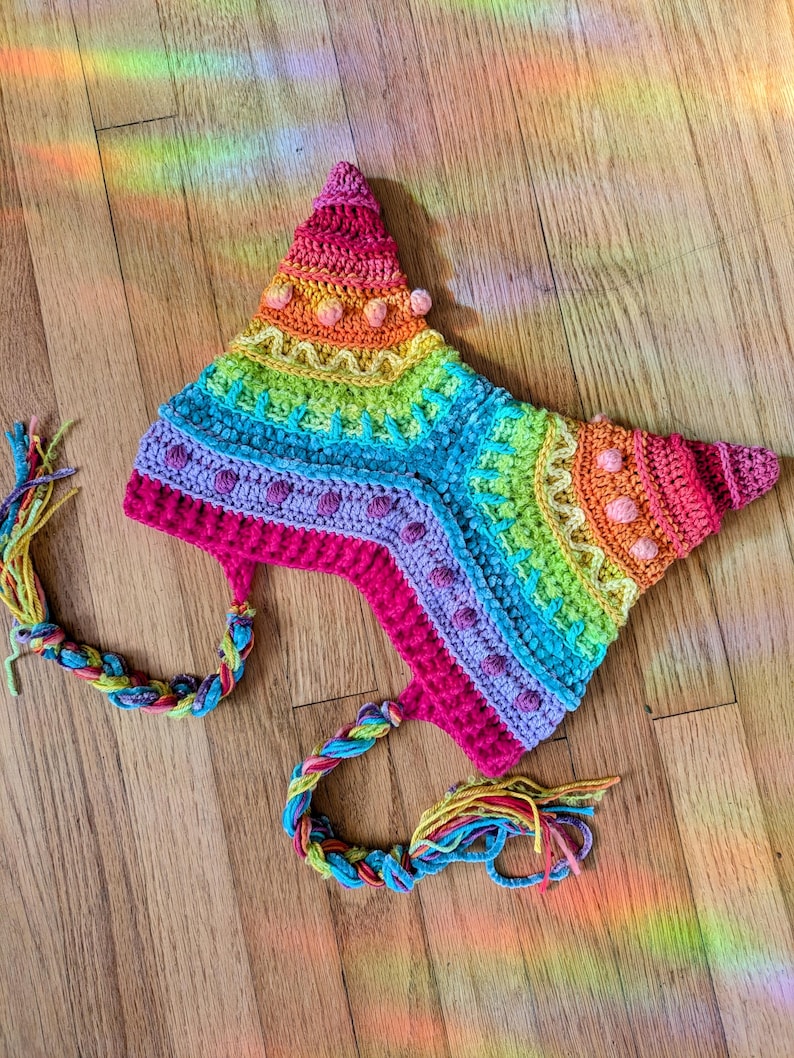 May include: A colorful, handmade crochet hat in a star shape. The hat features a rainbow of colors, including pink, orange, yellow, green, and blue. It has braided ties with colorful tassels. The hat is likely for a child or adult.