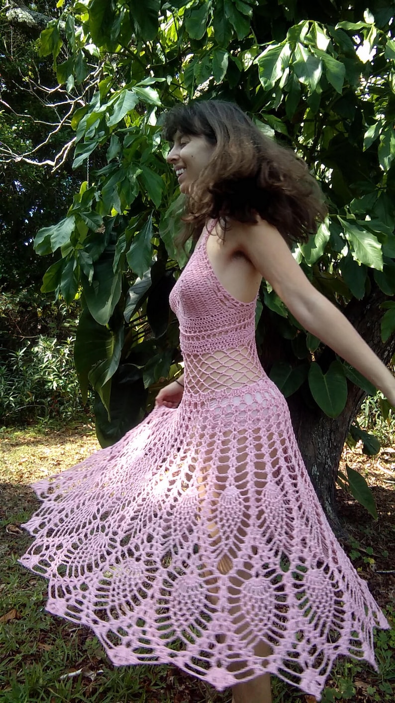 May include: A woman wearing a pink crocheted maxi dress with a delicate, lacy pattern. The dress has a halter neckline and a flowing skirt.