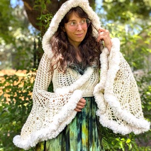 May include: A cream-colored crochet hooded shrug featuring an open-knit pattern, long flared sleeves, and a soft, fuzzy white trim along the hood and sleeve edges. The garment is worn over a green and black tie-dye dress with a pleated skirt.