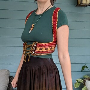 Puede incluir: Un chaleco de ganchillo con un esquema de color rojo, naranja y marr&oacute;n. El chaleco tiene un cierre de encaje en la parte delantera y se lleva sobre una camisa verde.