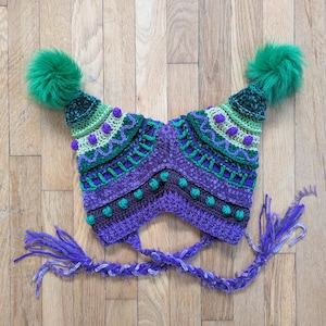 May include: A handmade, colorful crochet hat with a unique design. The hat features a combination of purple, green, and white yarn, with decorative bobbles and a braided tie. Two large green pom-poms adorn the top of the hat, adding a playful touch.
