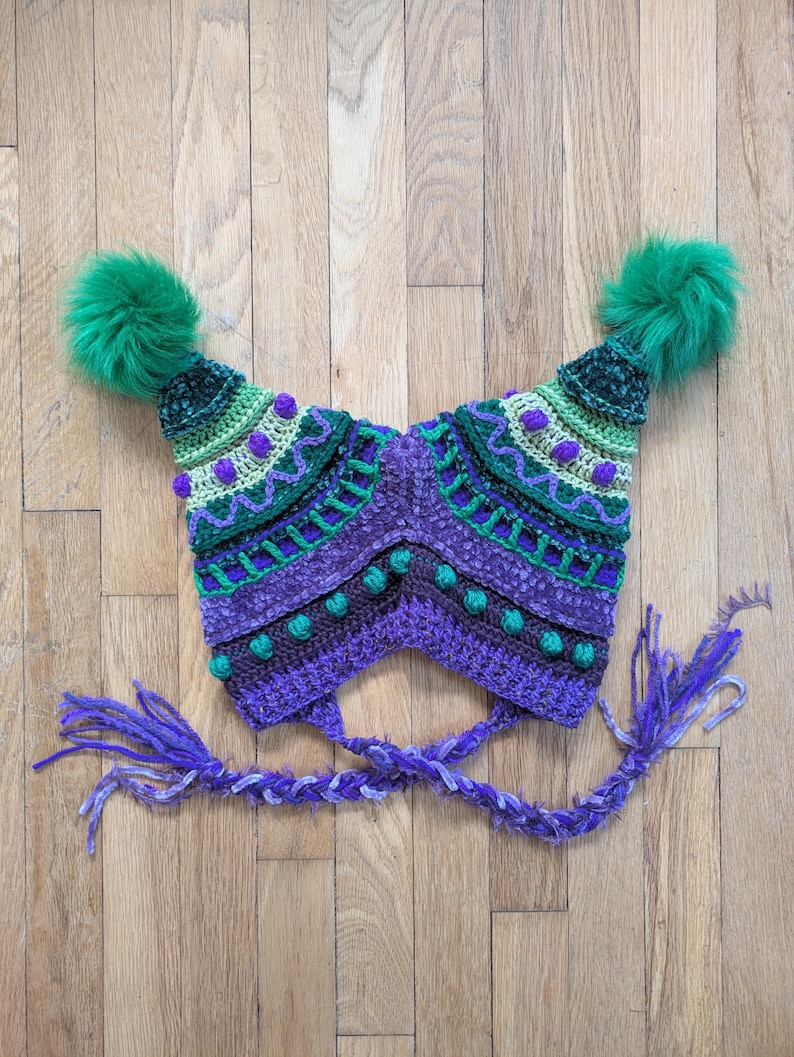 May include: A handmade, colorful crochet hat with a unique design. The hat features a combination of purple, green, and white yarn, with decorative bobbles and a braided tie. Two large green pom-poms adorn the top of the hat, adding a playful touch.