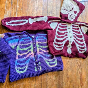 May include: Crocheted skeleton-themed sweaters in purple and burgundy. One purple sweater features a rainbow-colored skeleton design, while the burgundy sweater has a white skeleton design. A matching burgundy hood with a skull design is also present.