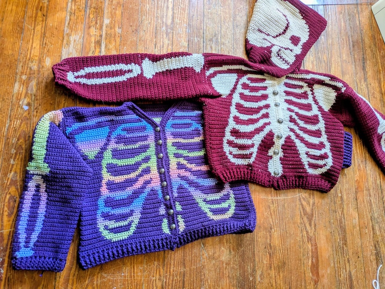 May include: Crocheted skeleton-themed sweaters in purple and burgundy. One purple sweater features a rainbow-colored skeleton design, while the burgundy sweater has a white skeleton design. A matching burgundy hood with a skull design is also present.