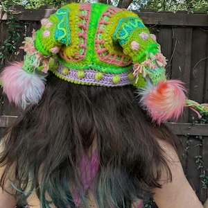 May include: A vibrant, handmade crochet hat in shades of neon green, pink, and turquoise. The hat features a whimsical design with fluffy pink pom-poms and decorative accents. The hat is a unique, eye-catching accessory.