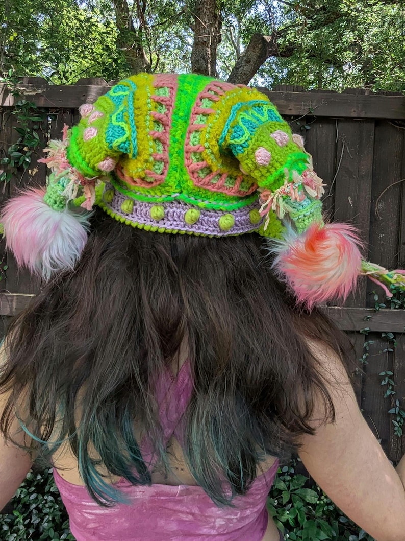 May include: A vibrant, handmade crochet hat in shades of neon green, pink, and turquoise. The hat features a whimsical design with fluffy pink pom-poms and decorative accents. The hat is a unique, eye-catching accessory.