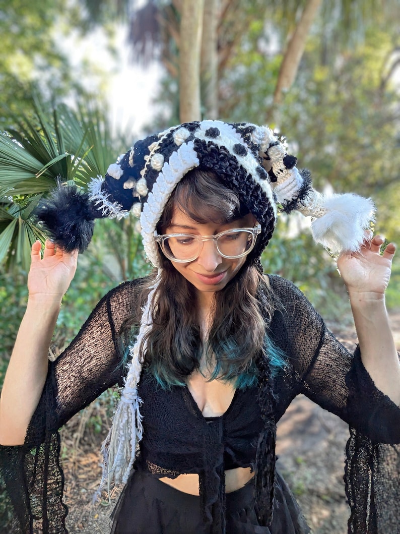 May include: A handmade, whimsical hat with a black and white polka dot pattern, white trim, and fluffy black and white pom-poms. The hat is a unique accessory, perfect for adding a touch of fun to any outfit.