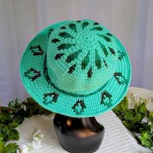May include: A turquoise crochet hat with a wide brim and a dark green leaf pattern. The hat is displayed on a black mannequin head against a white background with greenery.