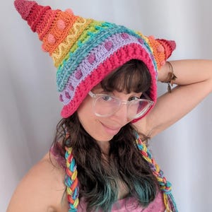 May include: A colorful, handmade crochet hat in a rainbow of hues, featuring a pointed top and textured details. The hat has a pink brim and is adorned with small, round accents. Braided hair extensions in matching colors complete the look.